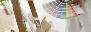 Handschuhe von Hausmaler Mann Dekorator wählen Sie die Farbe mit der Swatch-Probe, die Arbeit des Hauses zu renovieren, eine Holzleiter mit Pinsel und ein Eimer als Hintergrund, Nahaufnahme